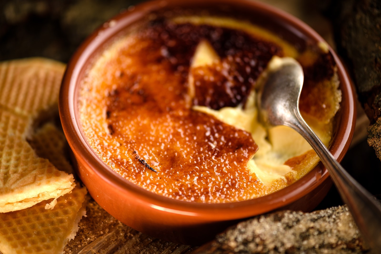 Crème brûlée in a terracotta ramekin with caramelized sugar top being cracked with a spoon, shortbread cookies alongside