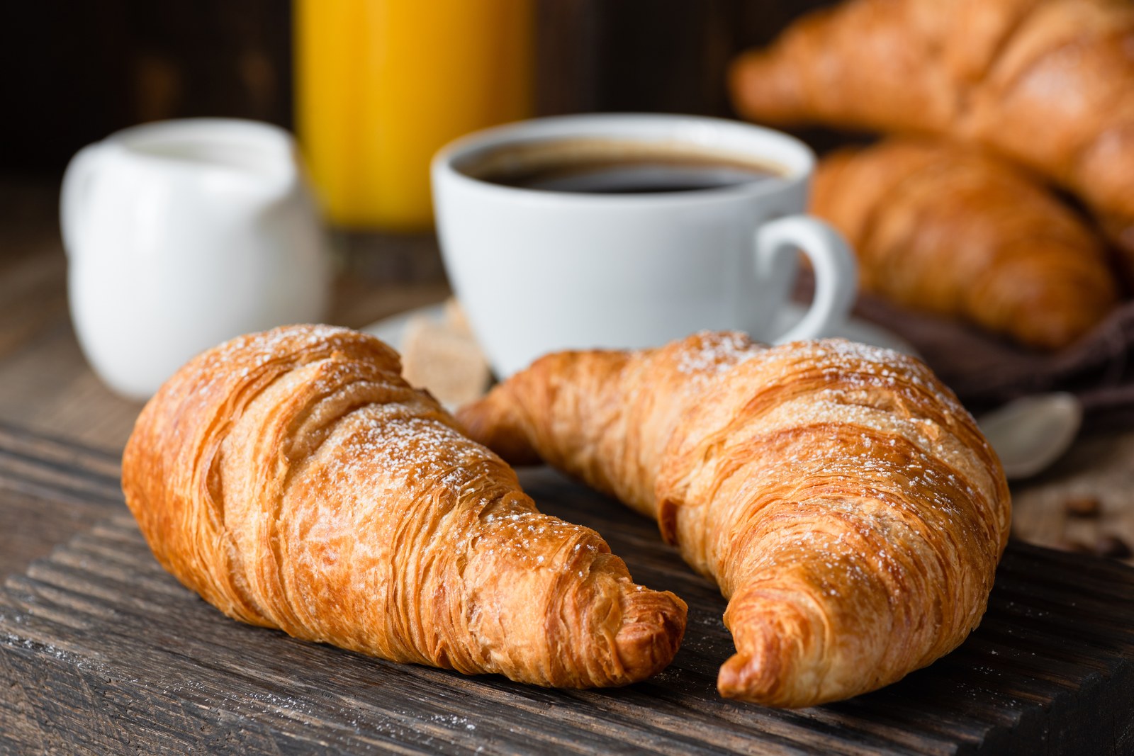 Two golden flaky croissants with visible laminated layers dusted with powdered sugar on a dark slate board, coffee cup in background
