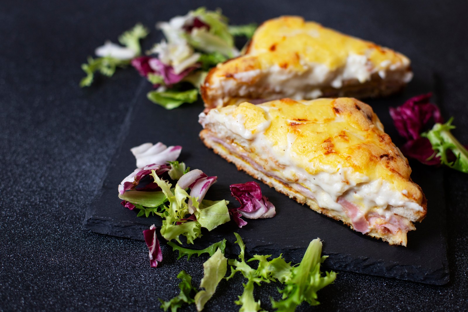 Two halves of croque monsieur on a dark slate plate showing melted cheese, ham layers, and béchamel sauce, garnished with mixed greens