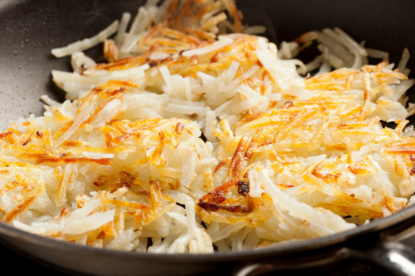 Crispy golden hash browns cooking in a skillet, showing shredded potatoes with browned edges and some white uncooked portions