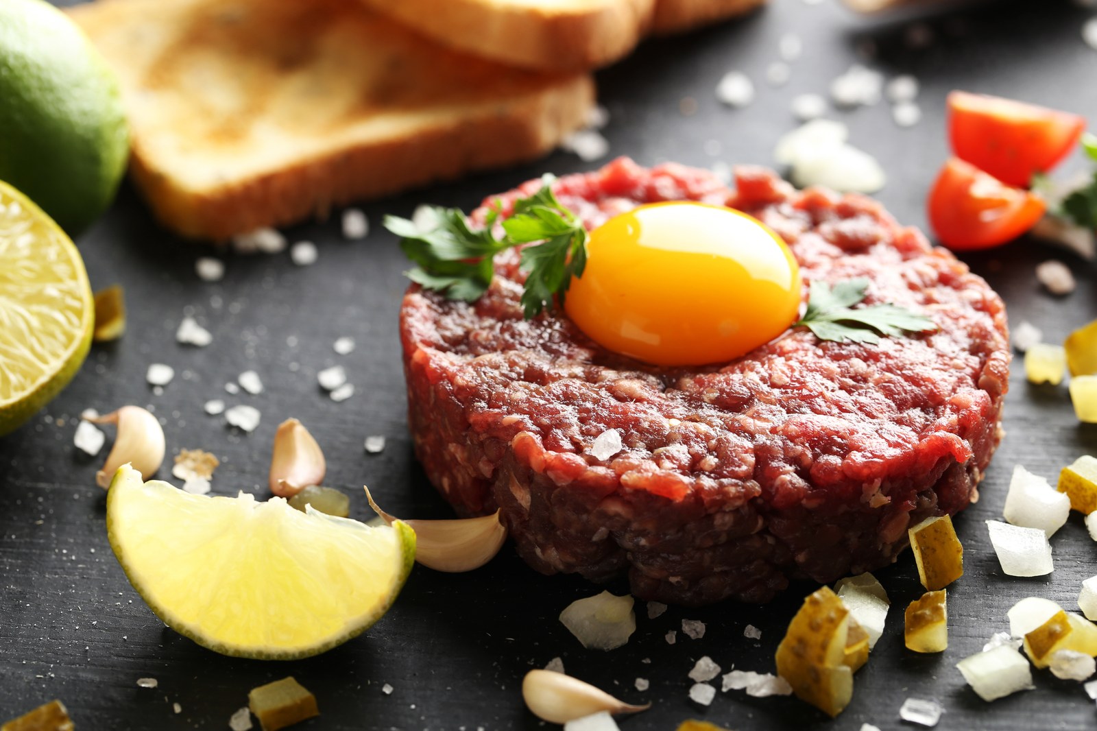 Classic French beef tartare with hand chopped raw beef seasoned with capers, onions, and parsley, formed into a round patty topped with a fresh egg yolk, garnished with parsley and surrounded by lemon wedges and minced garlic on a dark slate plate