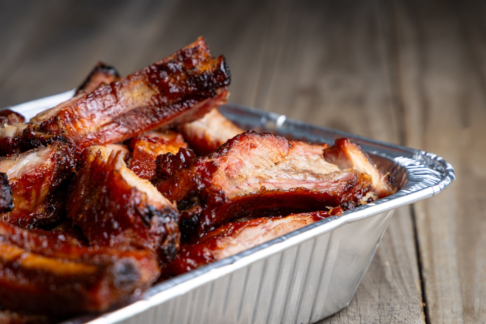 Braised bone-in beef short ribs with deep mahogany glaze, caramelized crust, charred edges, tender meat in aluminum pan