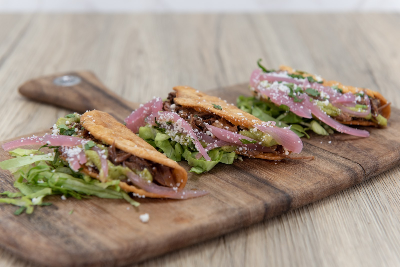 Braised short rib tacos on grilled corn tortillas topped with pickled red onions, fresh cilantro, cotija cheese crumbles on wooden cutting board
