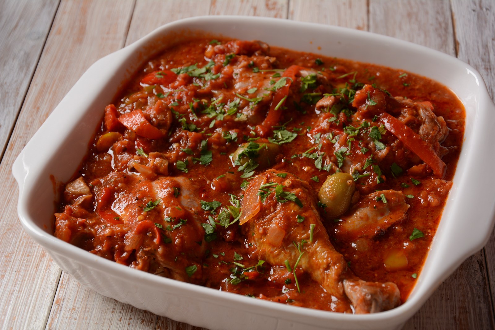 Chicken cacciatore in white baking dish with tomato sauce, chicken pieces, bell peppers, olives, fresh parsley garnish on rustic white wood background