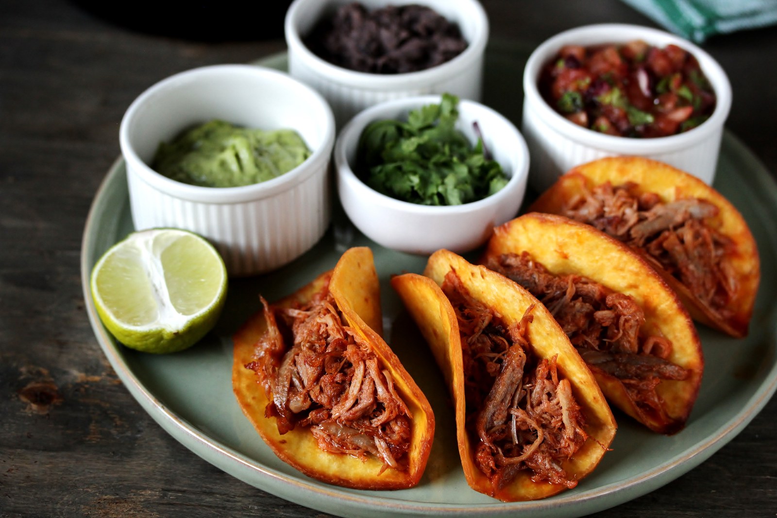 Four golden crispy taco shells filled with shredded chicken on a teal plate, accompanied by small white bowls of guacamole, cilantro, black beans, and salsa, with lime halves