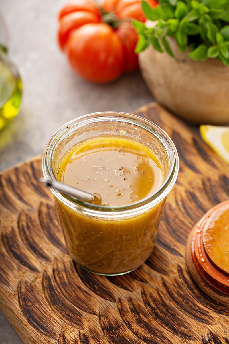 Homemade creamy Italian salad dressing in glass jar with visible herbs and spices, golden olive oil layer, fresh tomatoes and basil in background on wooden board