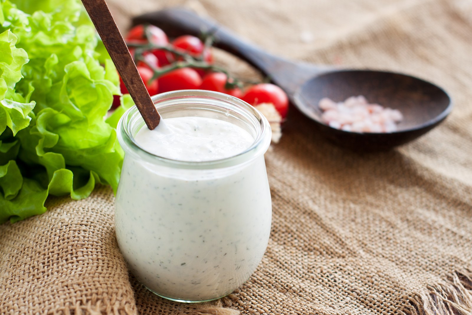 Creamy parmesan peppercorn dressing in glass jar with visible black pepper flecks, thick consistency, lettuce and cherry tomatoes in background on burlap cloth