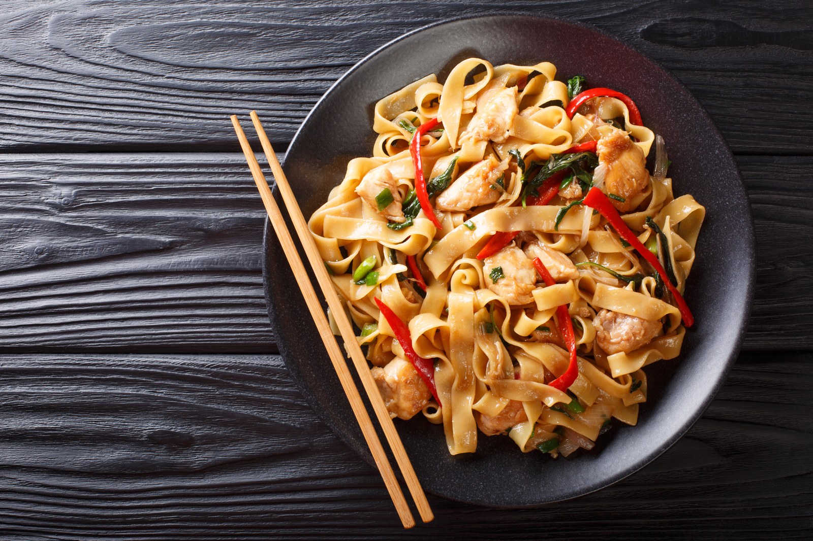 Plate of pad kee mao drunken noodles with wide rice noodles, chicken, Thai basil, and red peppers