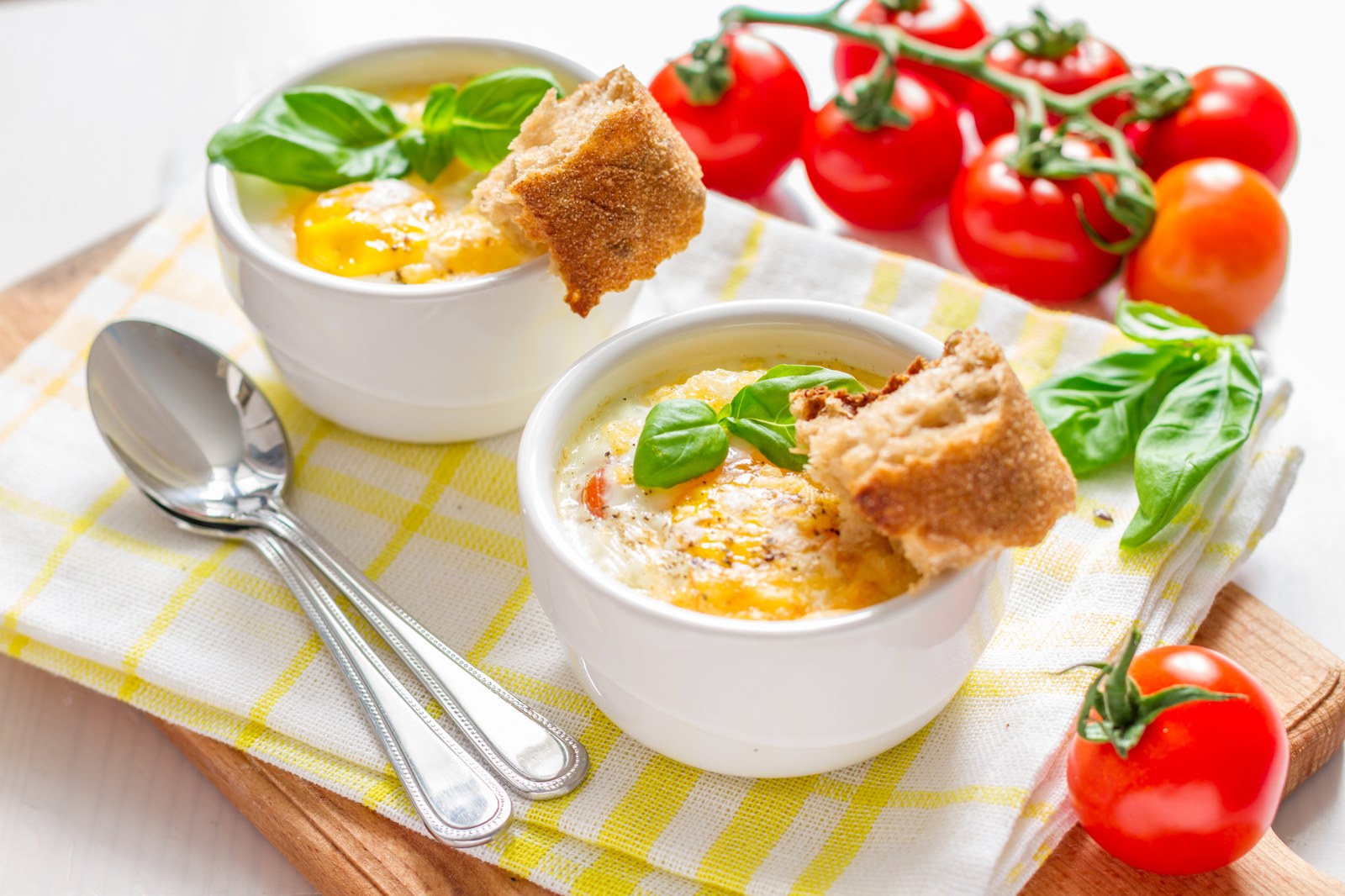 Two ramekins of eggs cocotte with baked eggs in cream, garnished with fresh basil and served with crusty bread