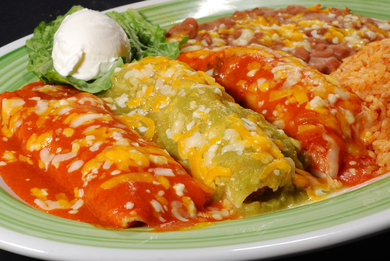 Plate of cheese enchiladas covered in red and green sauce with melted cheese, sour cream, and refried beans
