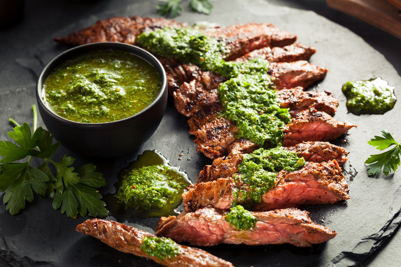 Sliced grilled flank steak topped with bright green chimichurri sauce, served on a dark slate platter