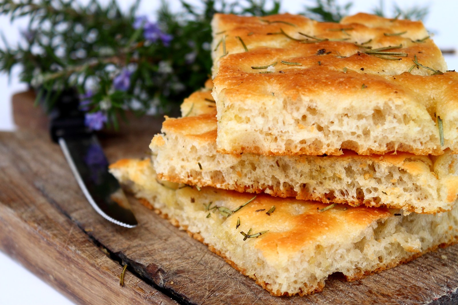 Sliced focaccia bread stacked on a wooden board, showing airy interior and golden top studded with fresh rosemary