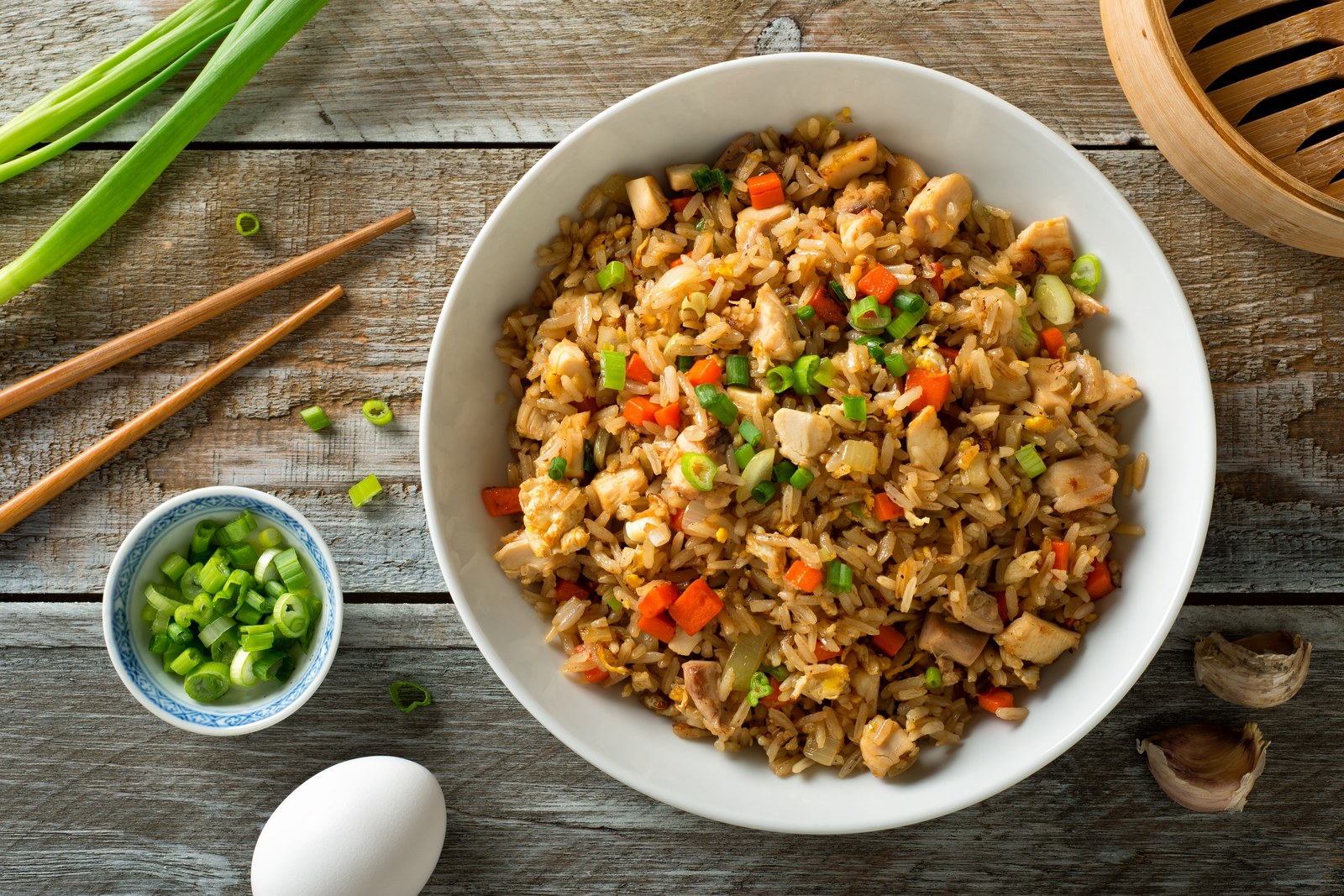Bowl of Asian style fried rice with scrambled eggs, diced chicken, carrots, and green onions, served with chopsticks on a wooden table