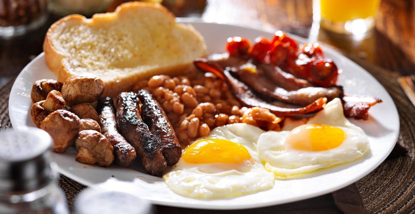 Traditional full English breakfast plate with two fried eggs, sausages, bacon, baked beans, mushrooms, grilled tomato, and buttered toast