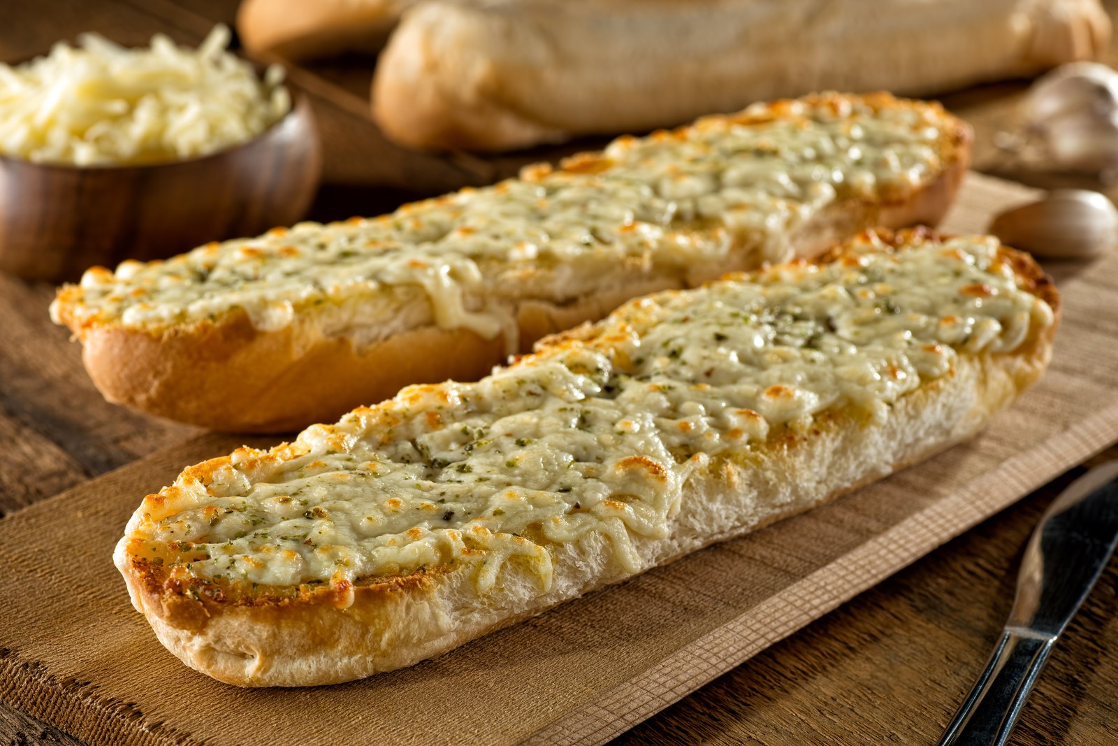 Three halves of garlic bread on a cutting board, topped with melted butter, minced garlic, herbs, and grated cheese