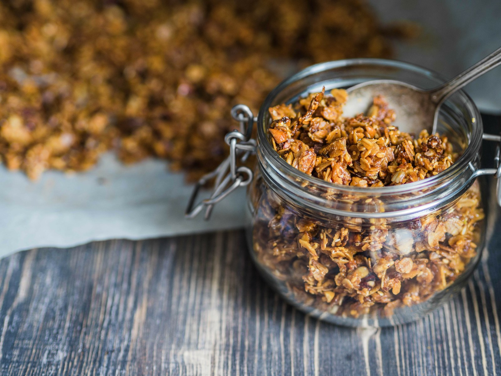 Homemade golden granola clusters in glass jar with visible oats, nuts, honey coating, crispy texture on dark wooden surface with parchment paper