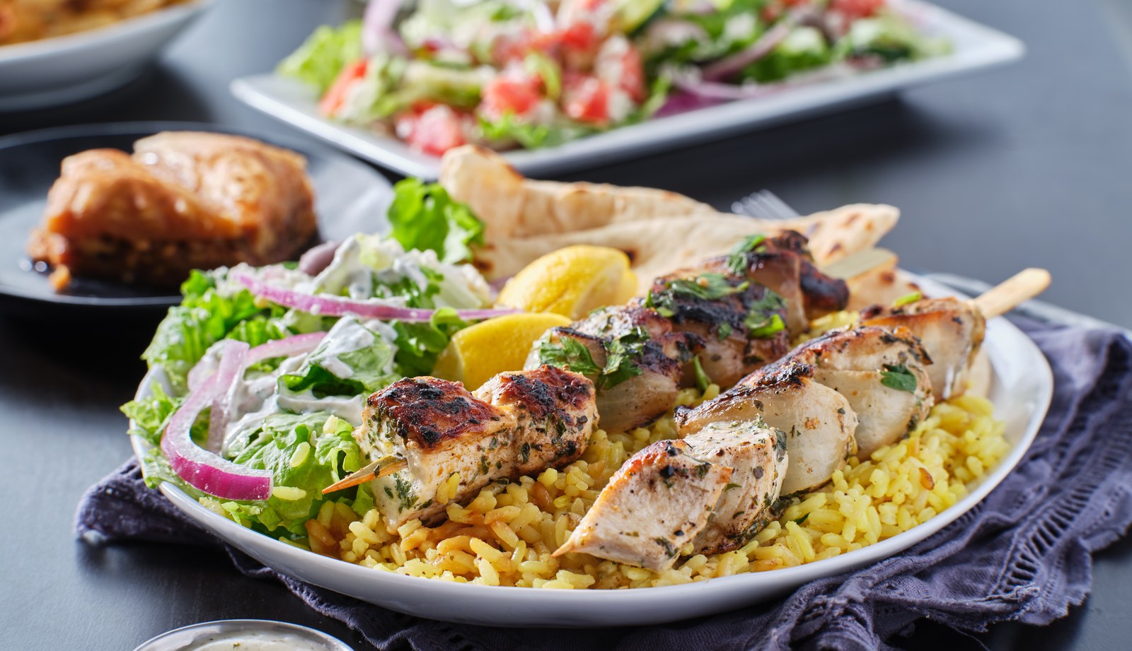 Plate of Greek chicken souvlaki skewers served over yellow rice with Greek salad, pita bread, and lemon wedges