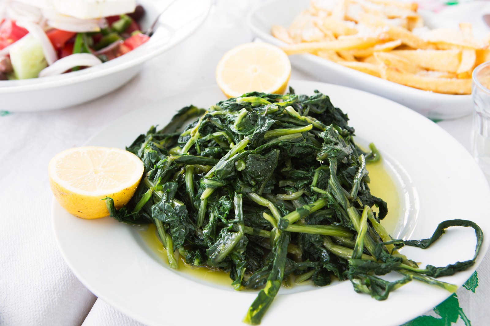 A plate of sautéed dark leafy greens garnished with lemon wedges and drizzled with olive oil, served alongside a Greek salad and fried potatoes