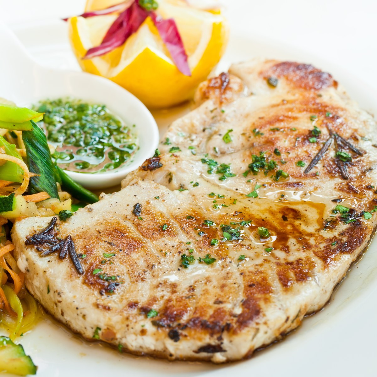 Two grilled swordfish steaks on a white plate with sautéed vegetables and a side of sauce
