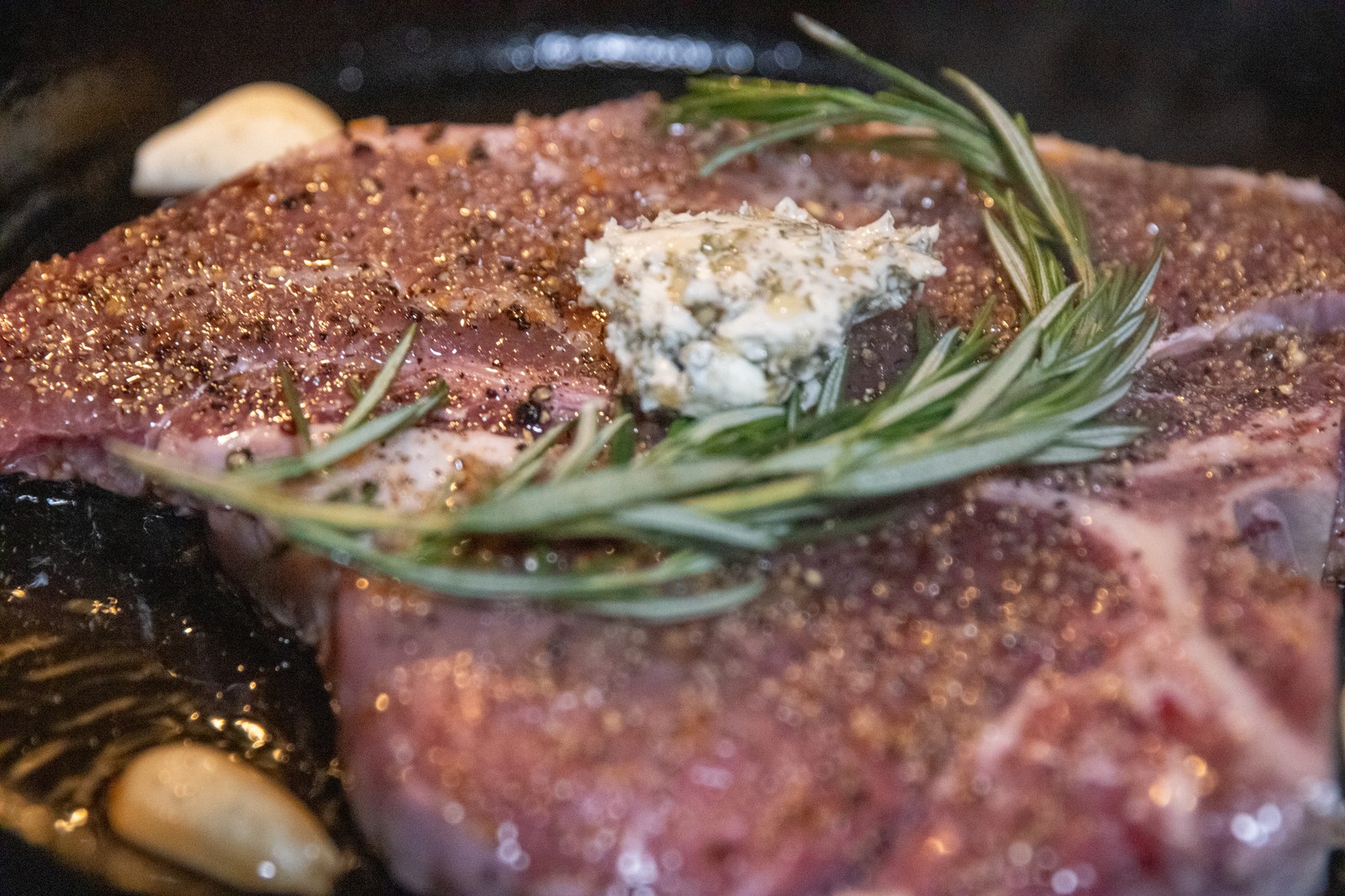 A raw hanger steak seasoned with salt, pepper, and rosemary, topped with herb butter in a cast iron skillet