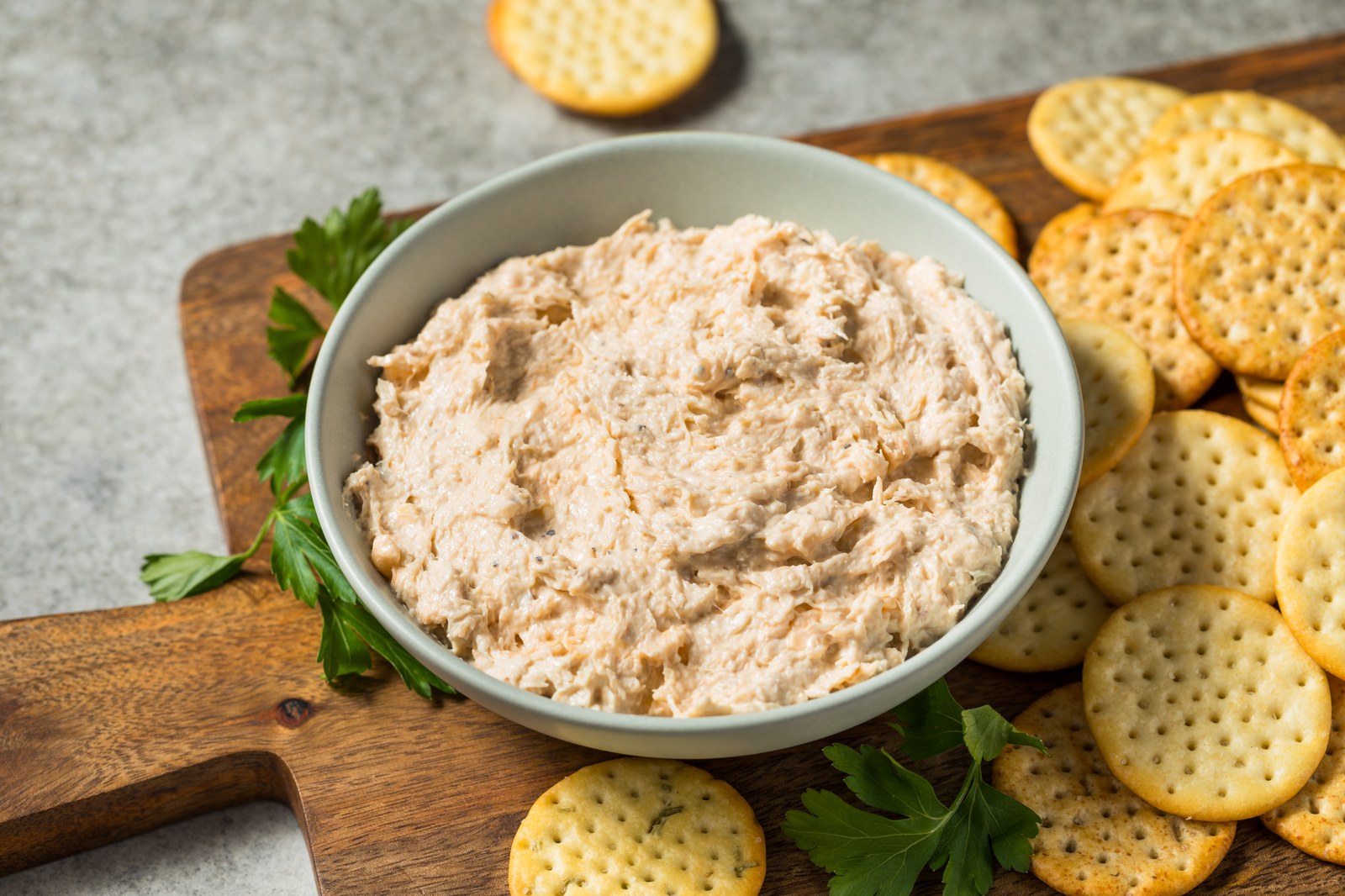 A white bowl filled with creamy beige crab dip surrounded by round crackers on a wooden cutting board
