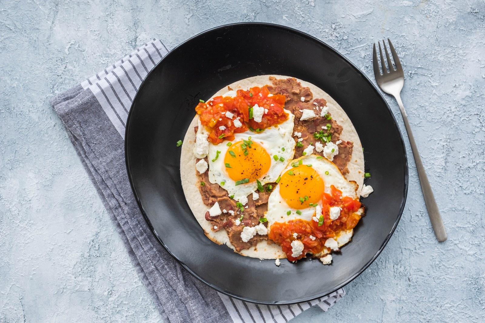 Two sunny side up eggs on a flour tortilla with refried beans, salsa, crumbled cheese, and fresh cilantro on a black plate