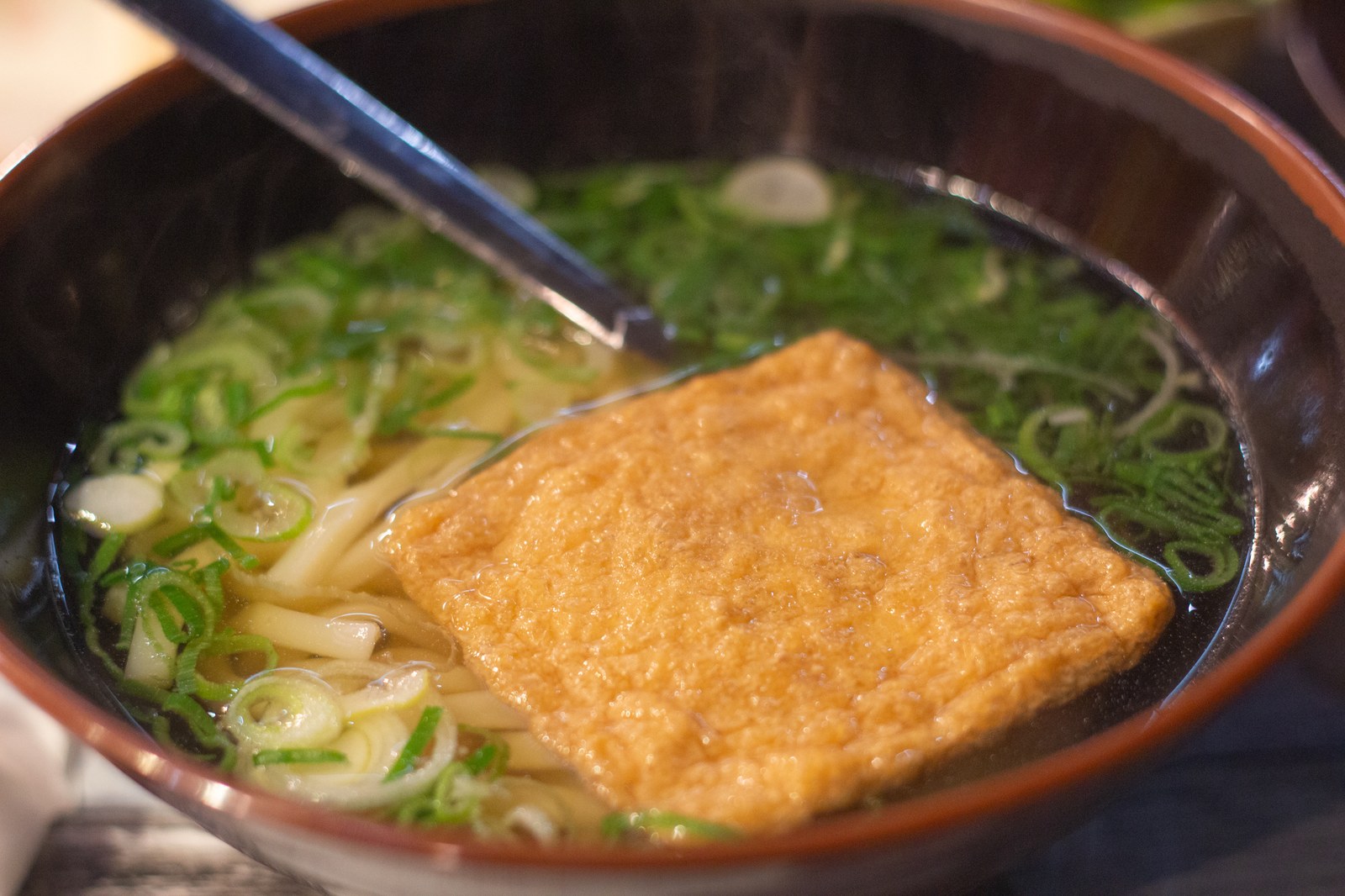 A bowl of udon noodle soup with a large piece of fried tofu and green onions in clear broth