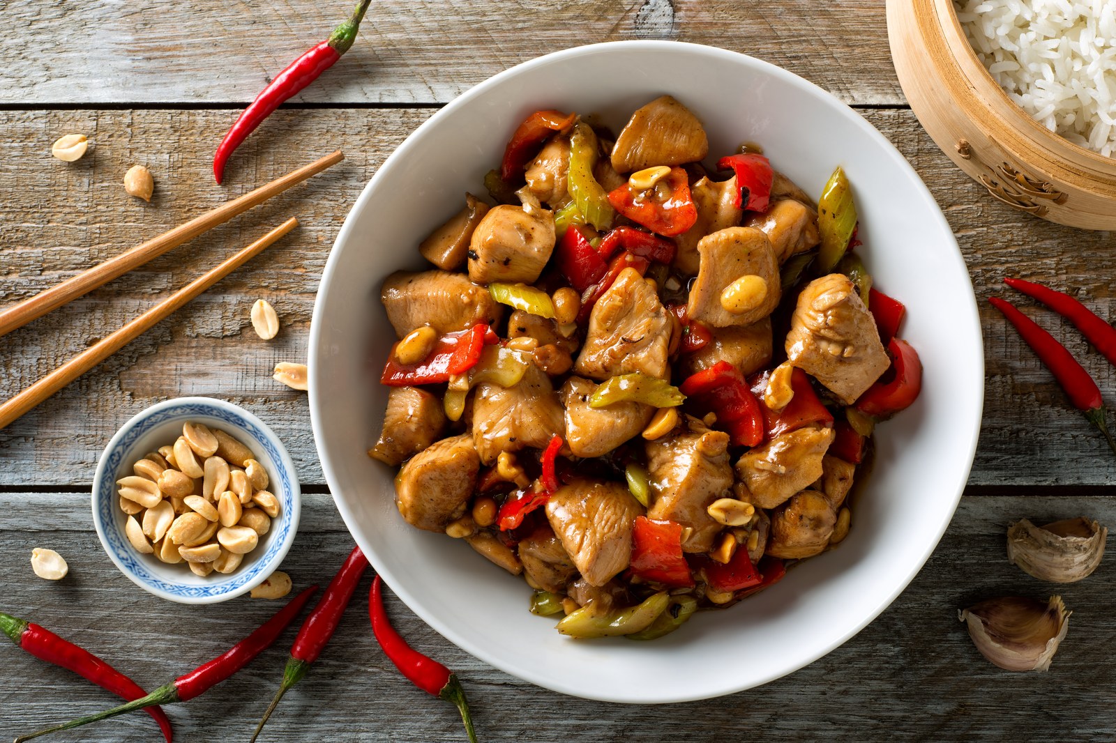 A white bowl of kung pao chicken with red and green peppers, peanuts, and chicken in glossy sauce with chopsticks and rice nearby