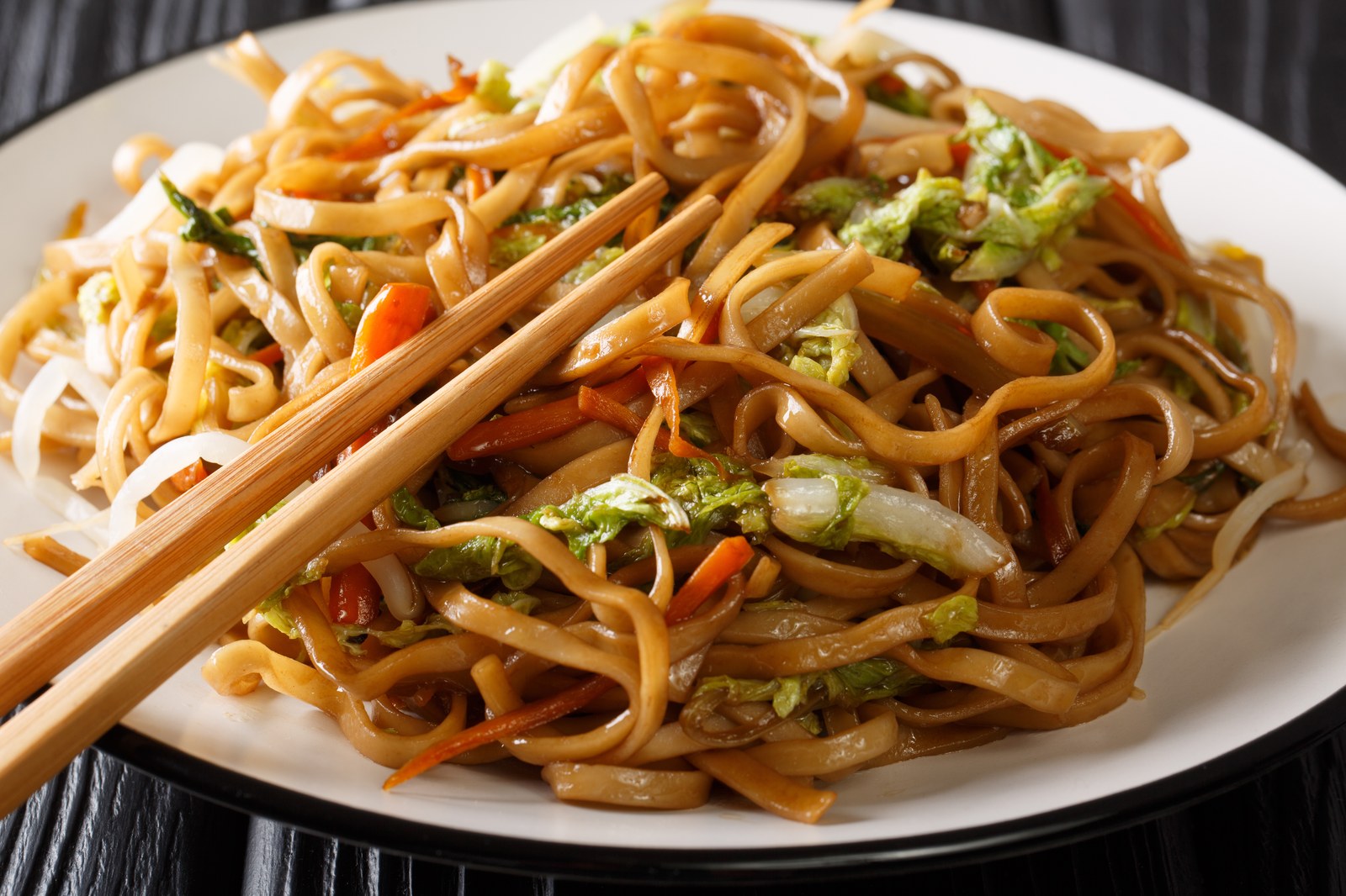 A white plate of stir fried lo mein noodles tossed with cabbage, carrots, and vegetables with chopsticks resting on top