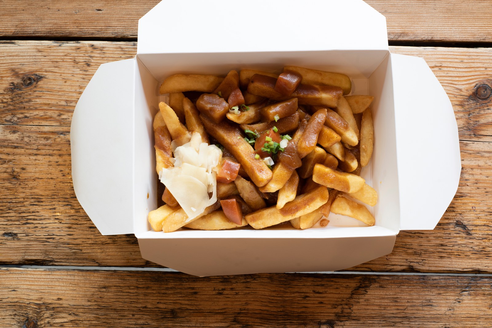 Loaded curry fries with golden french fries, brown curry gravy, white cheese curds, green onion garnish in white takeout box on wooden board