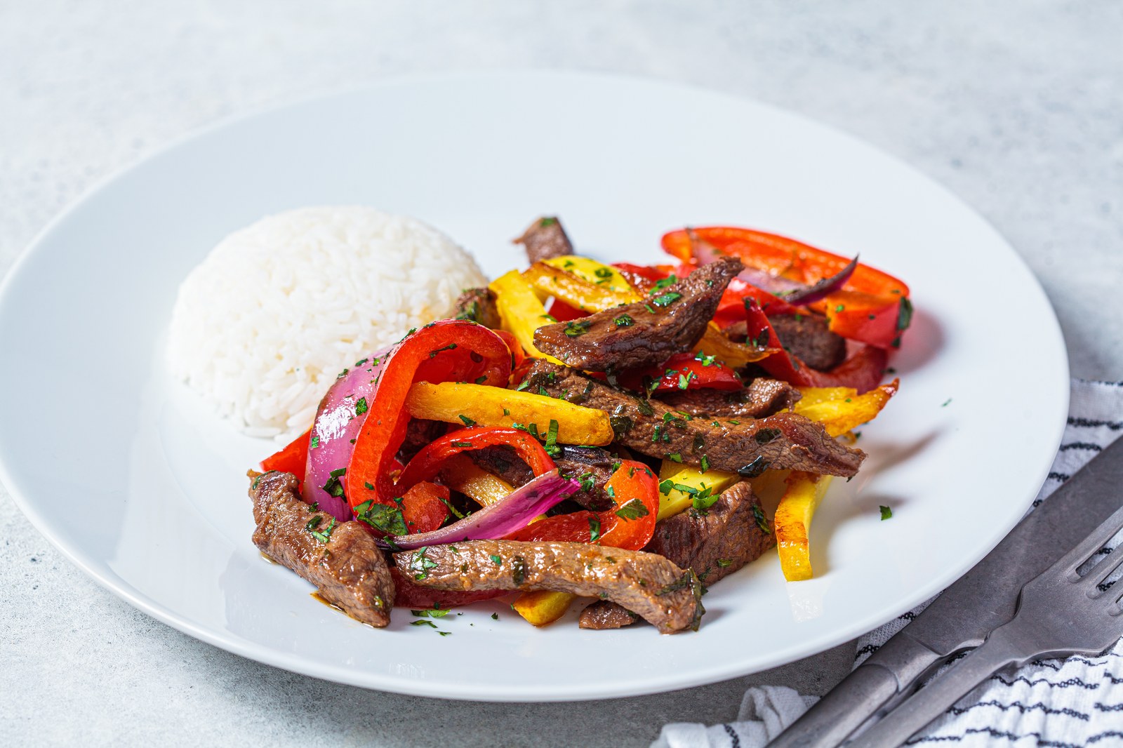 A white plate with Peruvian lomo saltado featuring sliced beef, red and yellow peppers, red onions, and white rice