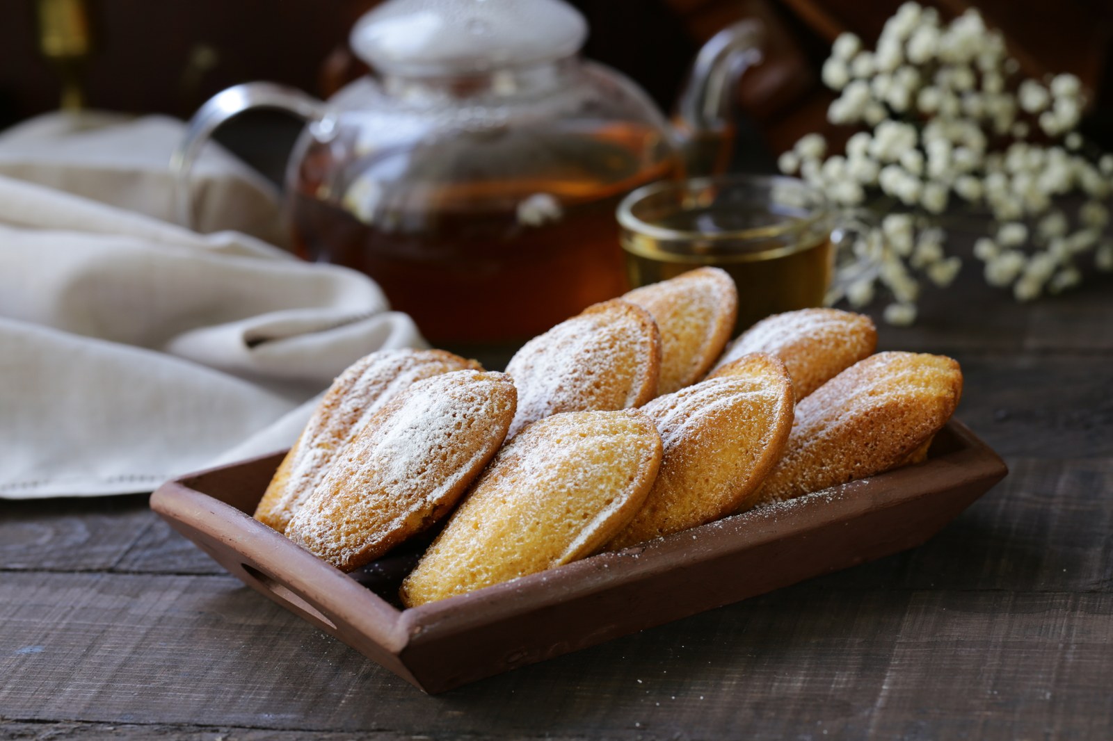 Shell shaped French madeleines dusted with powdered sugar arranged on a wooden tray with a teapot and flowers in the background