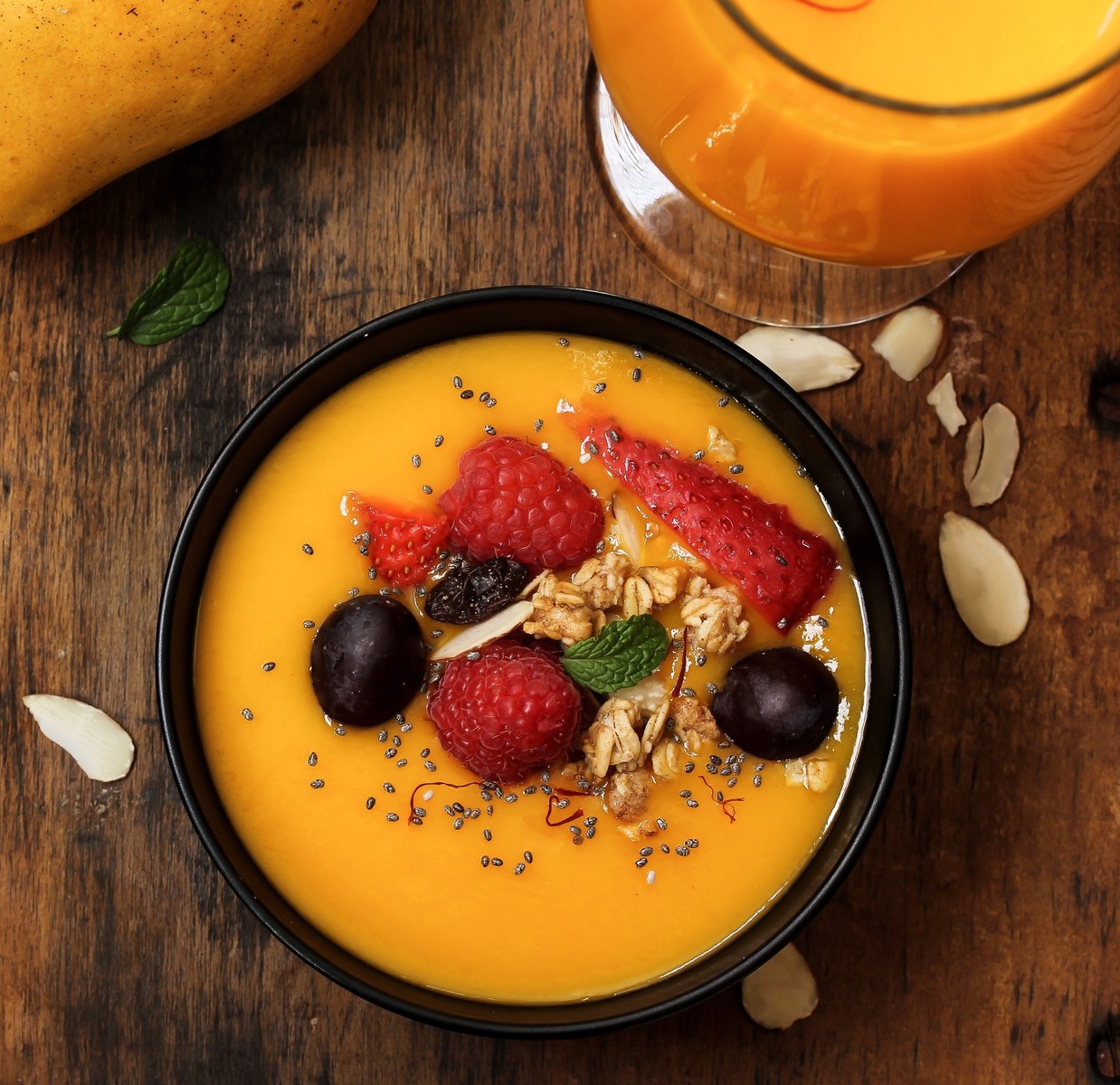 A black bowl containing vibrant orange mango smoothie topped with strawberries, blackberries, granola, chia seeds, and a mint leaf