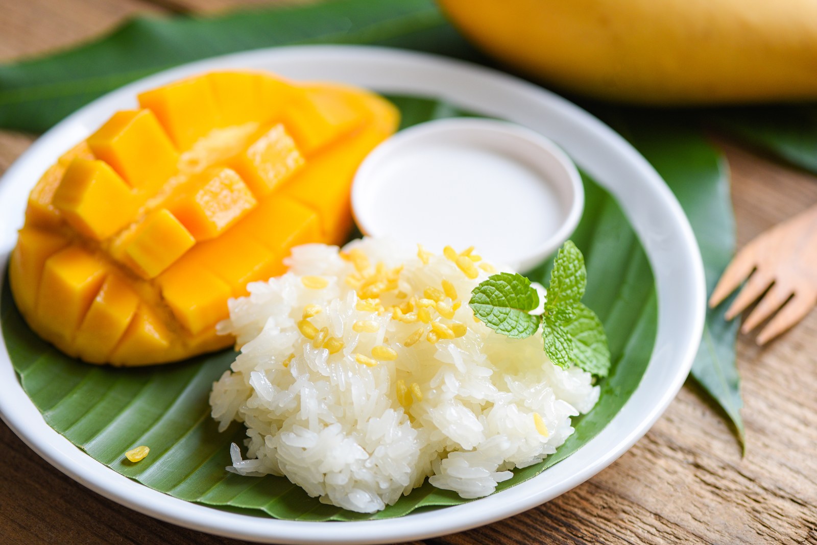 A white plate on a banana leaf with sweet sticky rice, fresh mango slices, and coconut cream garnished with mint
