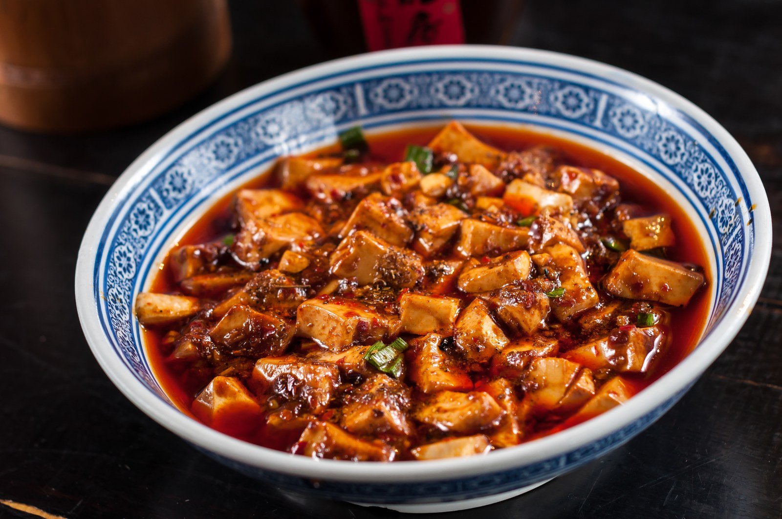 A blue and white patterned bowl filled with mapo tofu in spicy red sauce with ground meat and green onions
