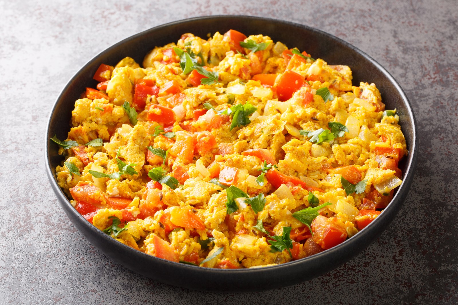 A black pan filled with scrambled eggs mixed with tomatoes, onions, and fresh herbs