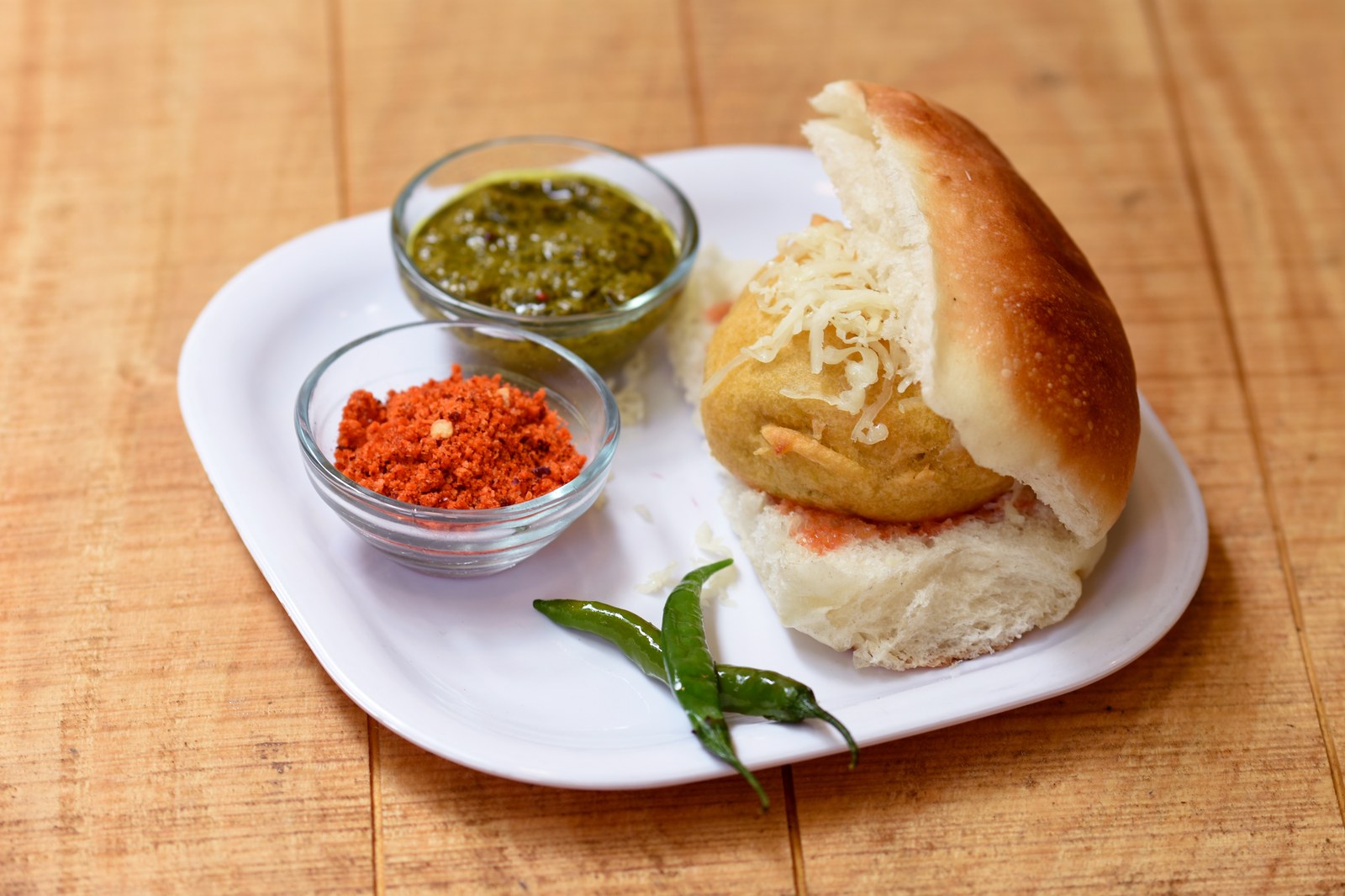 A burger bun filled with a spiced potato patty topped with shredded cheese and cilantro, served with green chutney and red chili powder