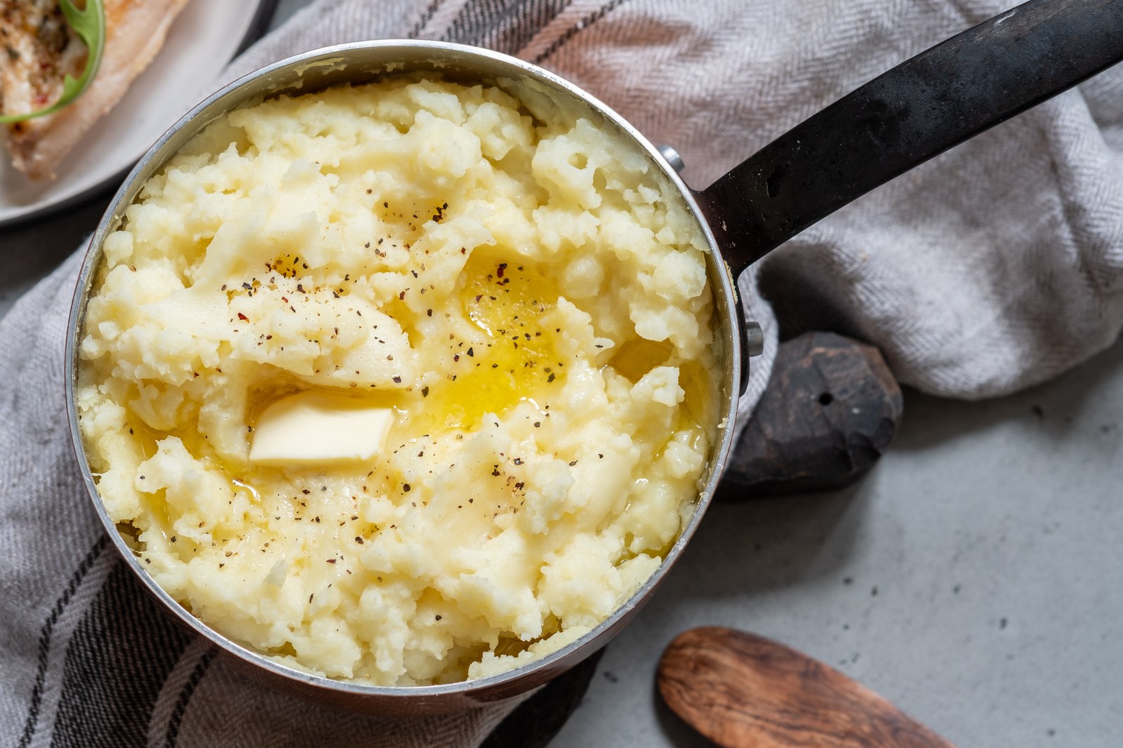 A silver pot of creamy mashed potatoes topped with a pat of butter and black pepper with a black handle