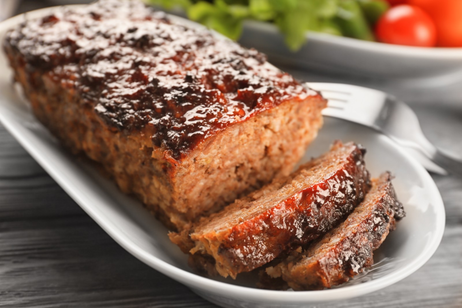 A sliced meatloaf with glossy glaze on top on a white platter with a salad in the background