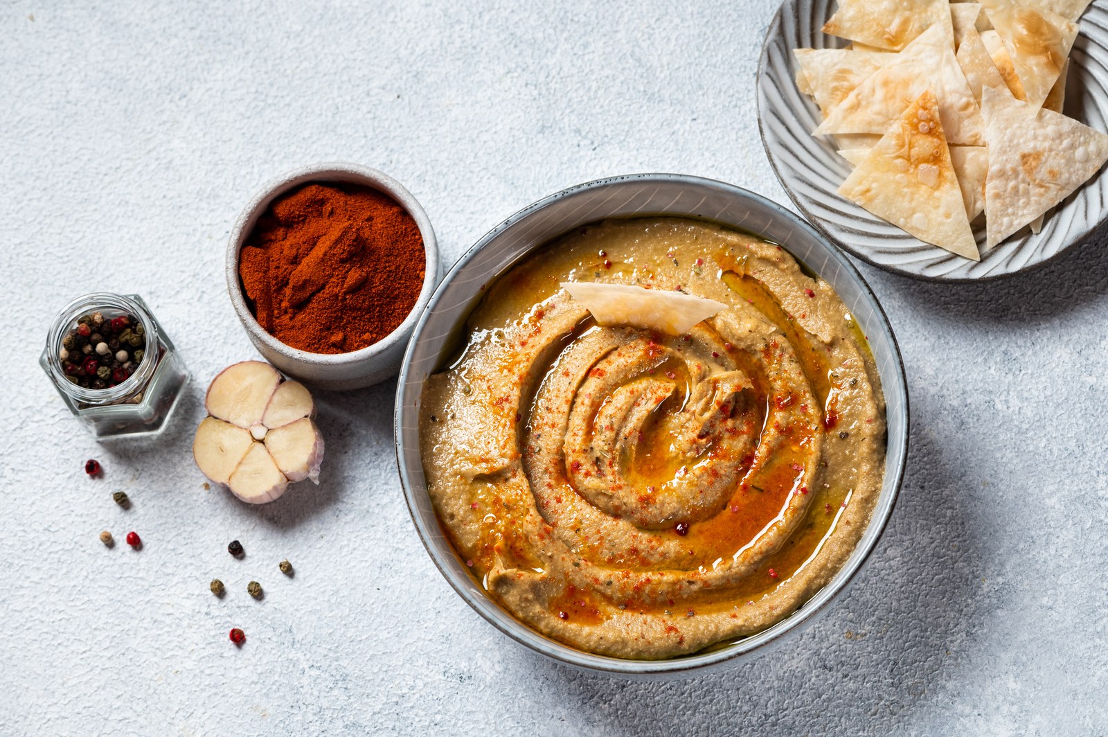 A bowl of creamy eggplant dip drizzled with olive oil and garnished with paprika, served with pita chips, garlic, paprika powder, and scattered peppercorns