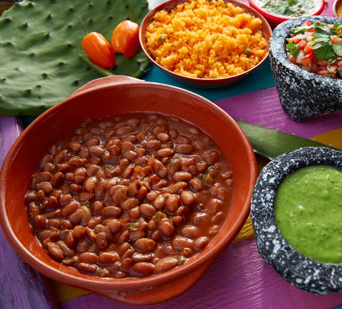 Terracotta bowl of stewed pinto beans surrounded by rice, salsa, and guacamole