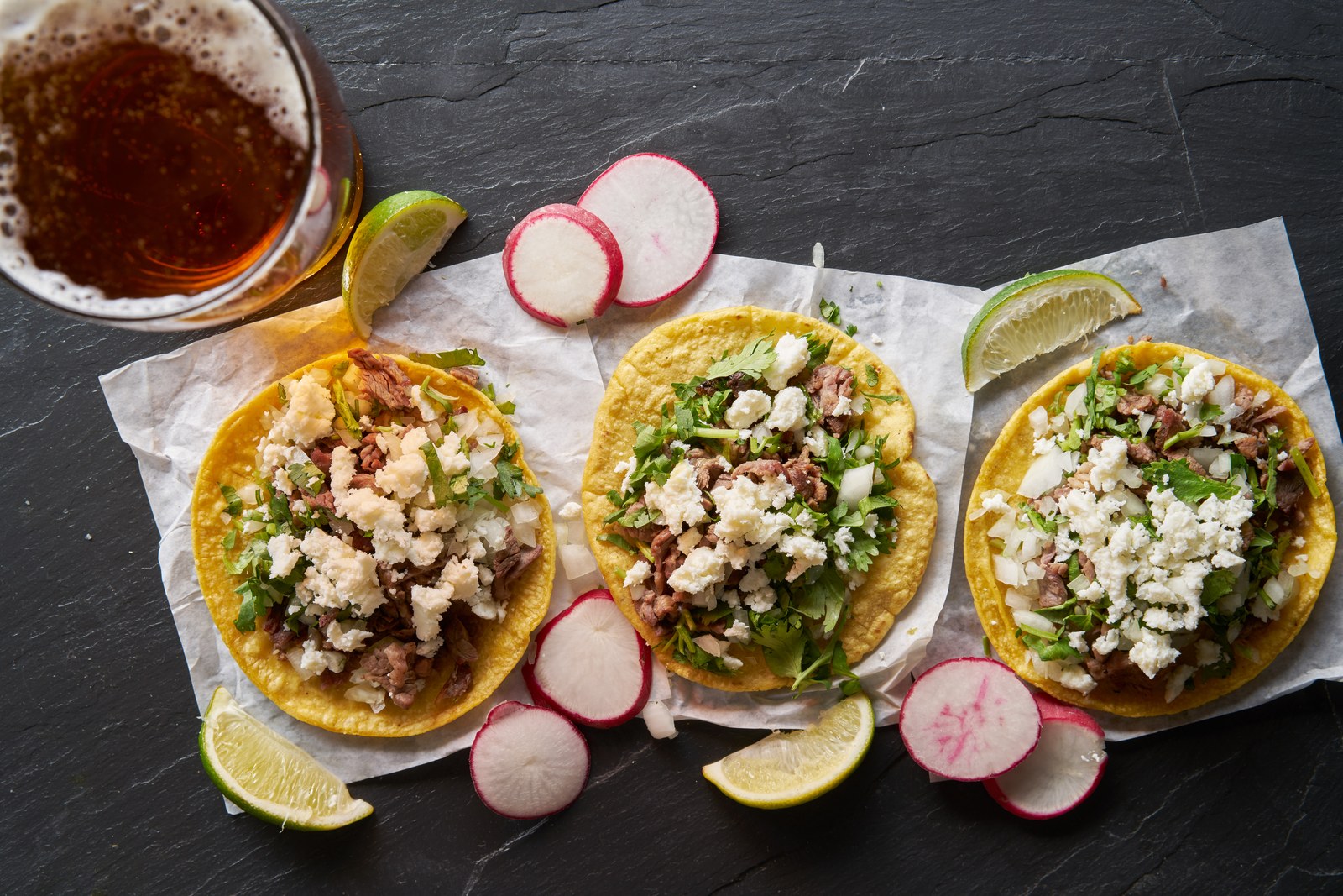 Migas tacos with chorizo, scrambled eggs, crispy tortilla strips, crumbled cotija cheese, cilantro, radish slices, lime wedges on dark slate with salsa