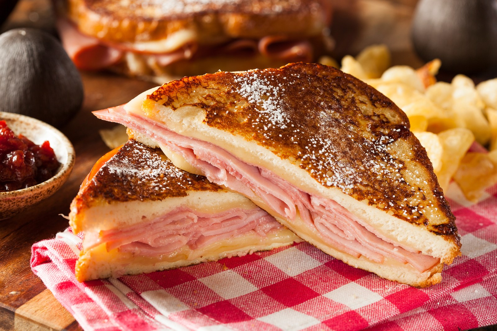 Monte Cristo sandwich with golden-brown french toast exterior, layers of sliced ham and melted cheese, powdered sugar dusting, raspberry jam and potato chips on red checkered cloth