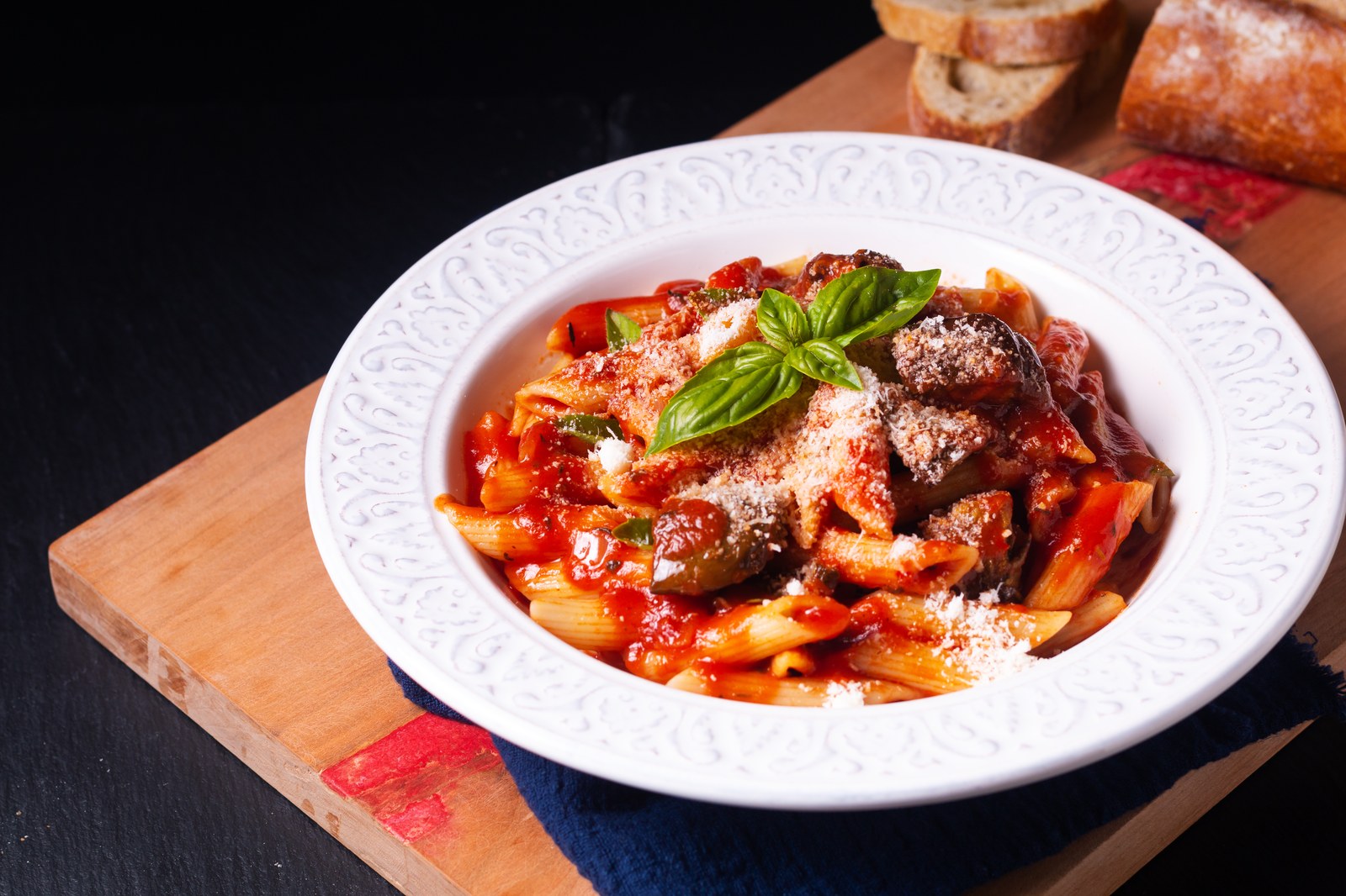 Pasta alla norma with penne, roasted eggplant chunks, tomato sauce, fresh basil leaves, grated ricotta salata cheese on white plate with Italian bread in background