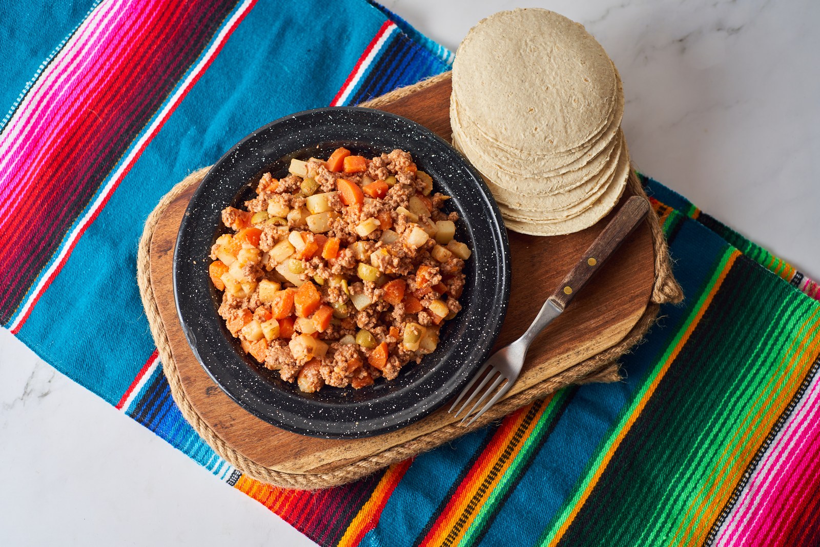Ground beef and vegetable hash served on a black plate with corn tortillas on a colorful textile
