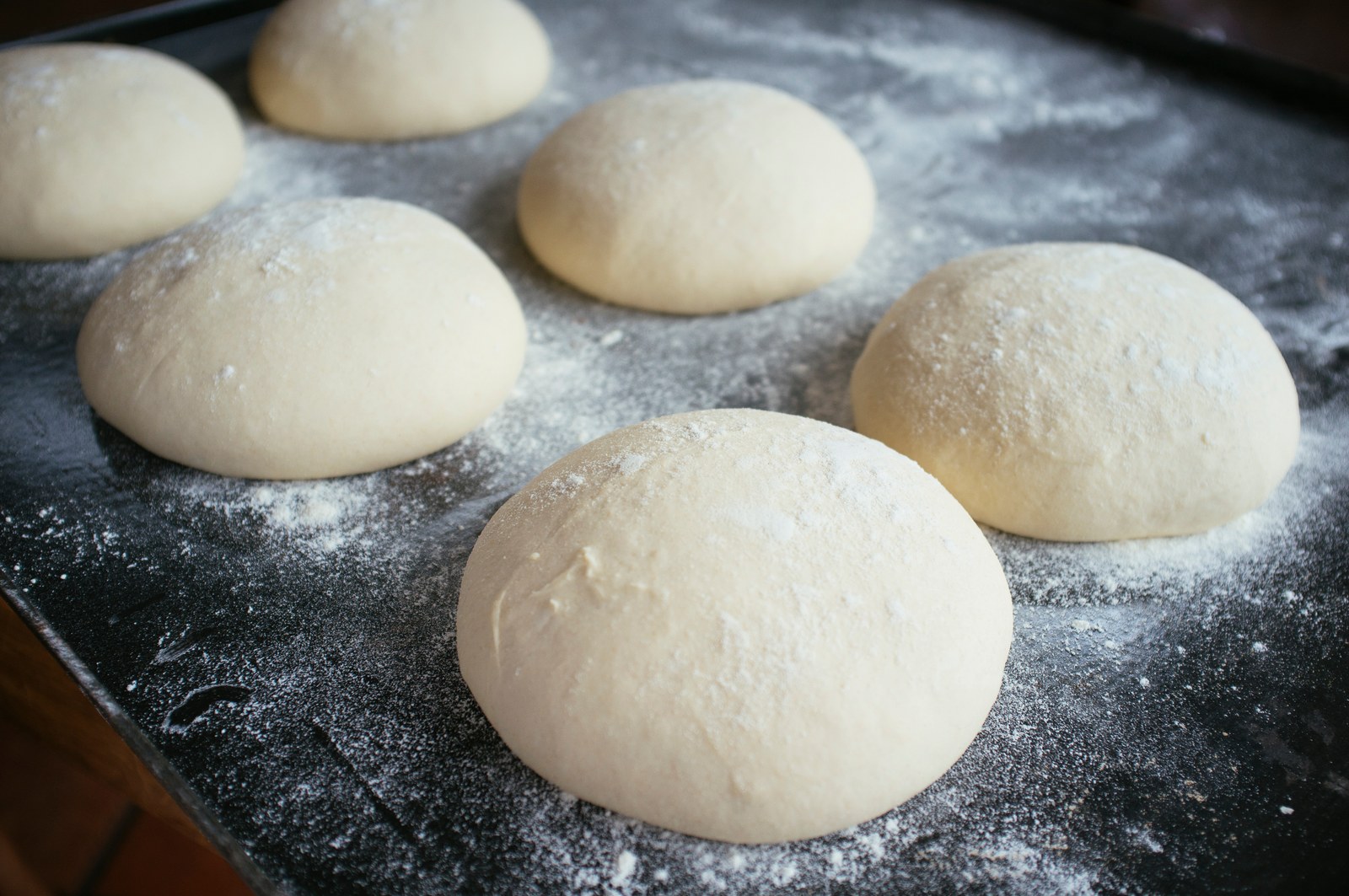 Six smooth balls of risen pizza dough dusted with flour on a dark baking sheet