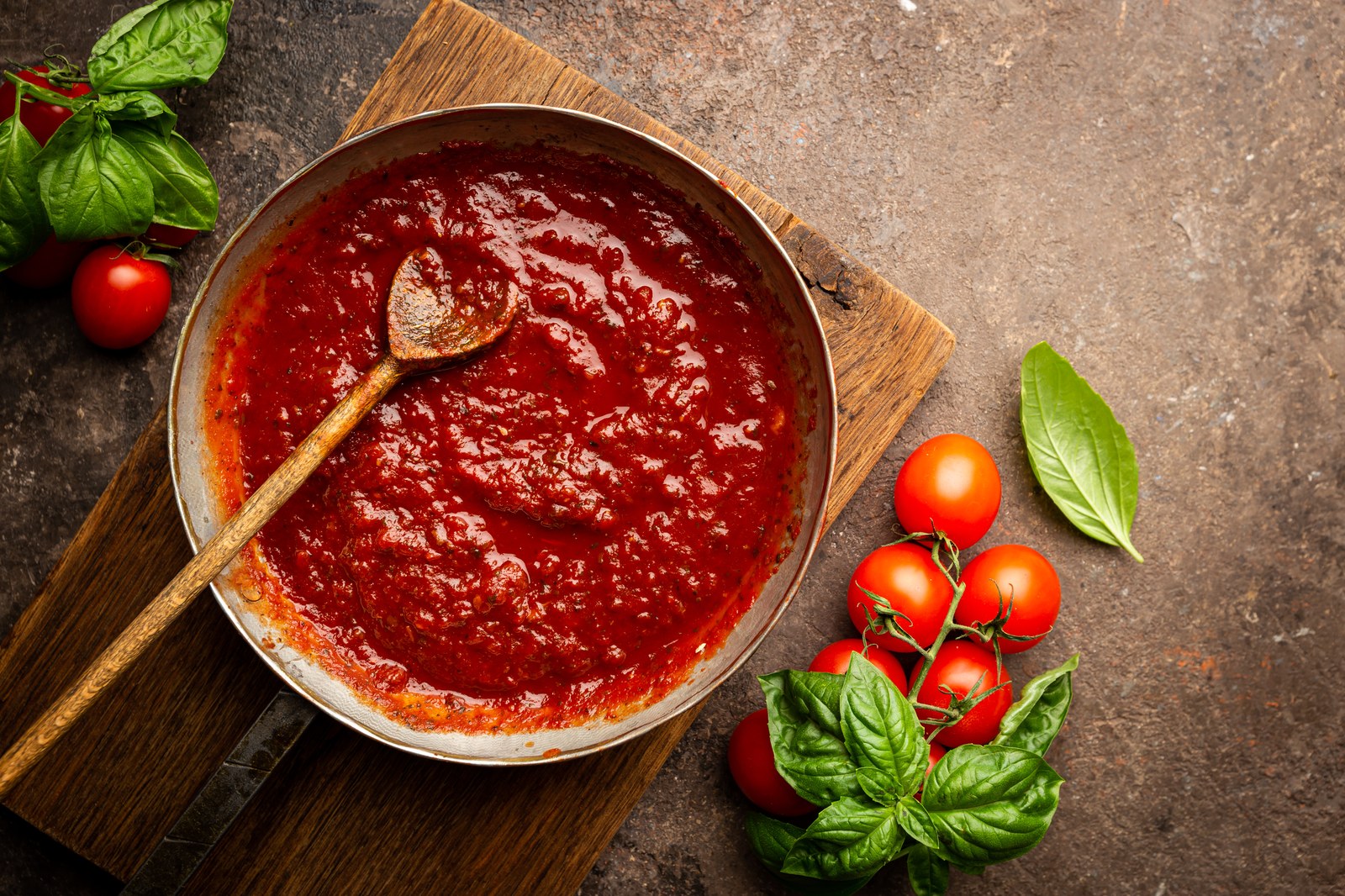 Pan of simmering red tomato pizza sauce with a wooden spoon and fresh basil and tomatoes nearby