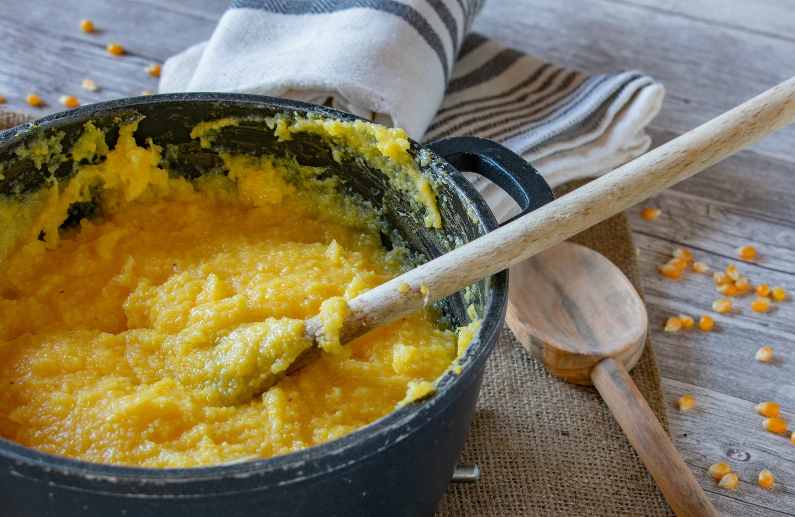 Creamy yellow polenta in a cast iron pot with a wooden spoon and scattered cornmeal