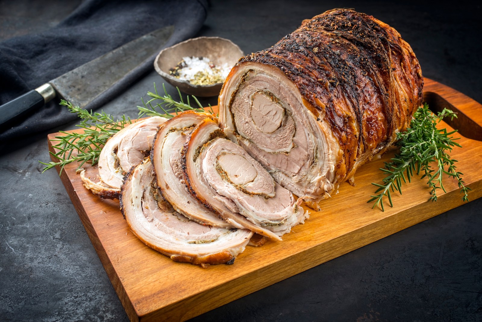 Rolled and roasted porchetta on a cutting board, sliced to show spiral of seasoned pork and crispy skin