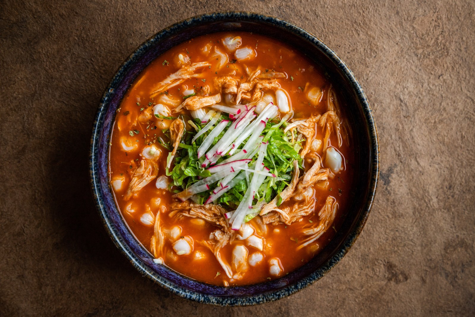 Bowl of red pozole soup with hominy, shredded chicken, and fresh garnishes of cabbage and radish